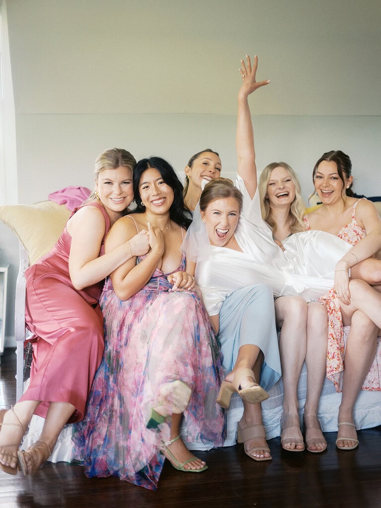 A group of six women joyfully posing together on a sofa, wearing colorful dresses, with one woman raising her hand in celebration.