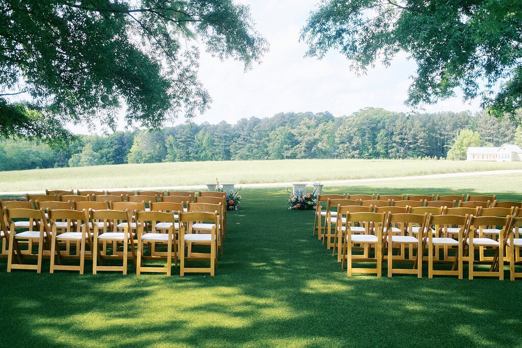 An outdoor wedding ceremony setup with wooden chairs arranged in rows on a green lawn, framed by trees, overlooking a field.
