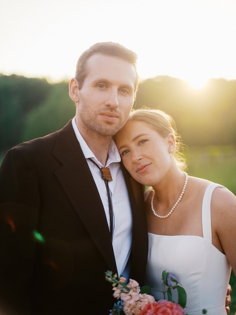 A bride and groom pose together during golden hour, with a soft sunlight illuminating their faces and the surrounding meadow.