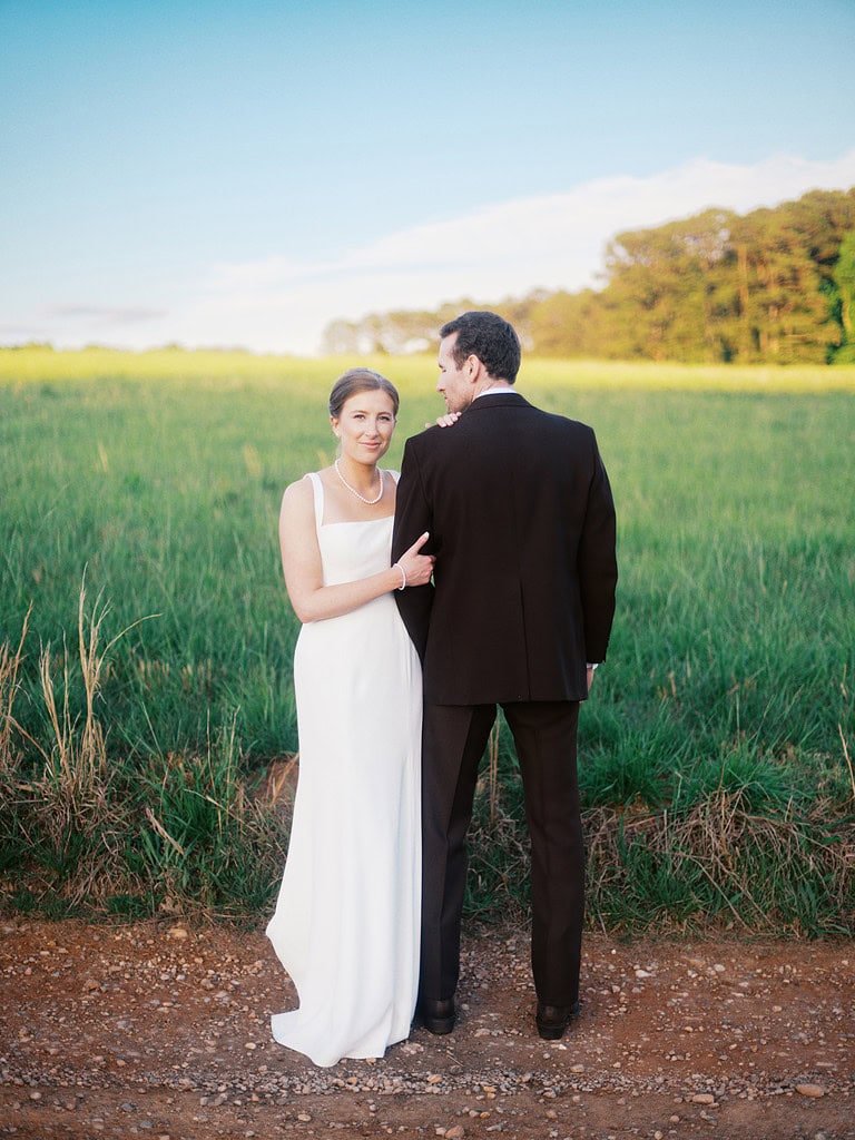 A couple standing together in a lush green field, facing away from the camera with sunlight illuminating their figures. The woman is wearing a white wedding dress, and the man is in a black suit.