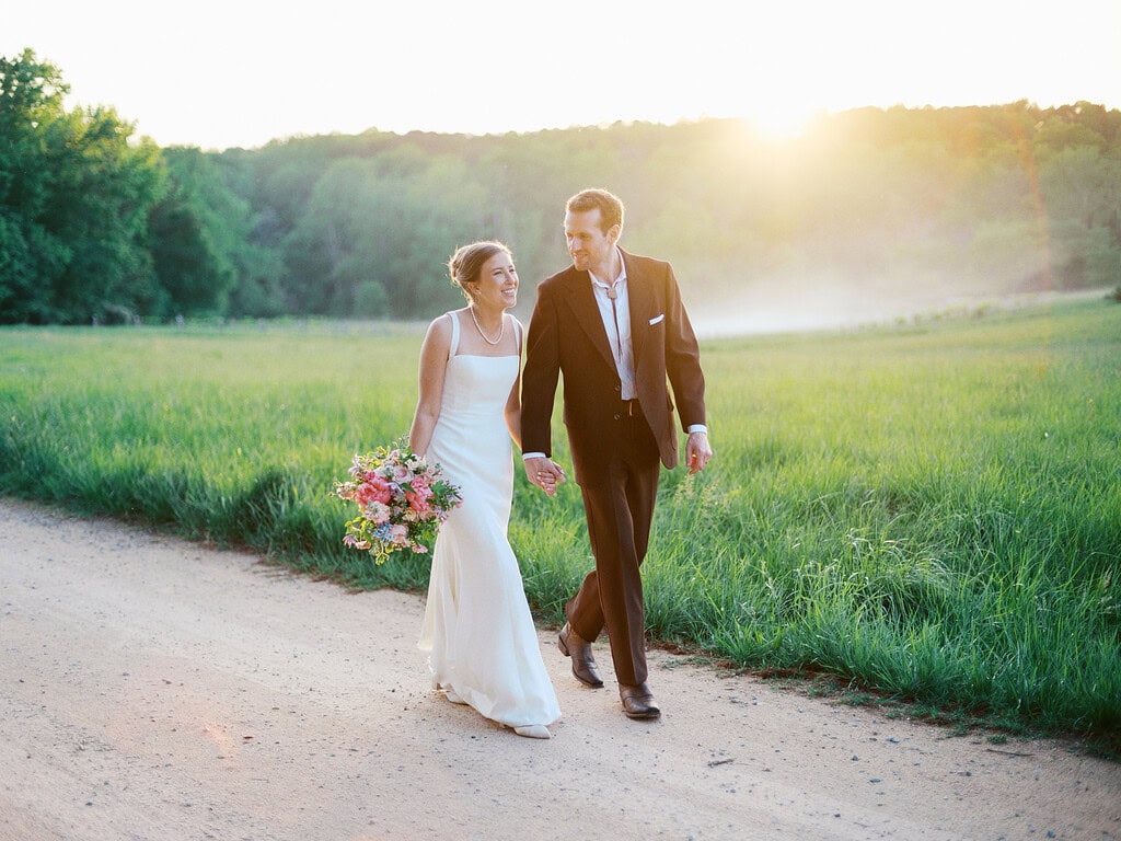 A bride and groom walk hand in hand at the Meadows at Firefly Farm down a dirt path surrounded by lush green fields, with the sun setting in the background, creating a warm glow.