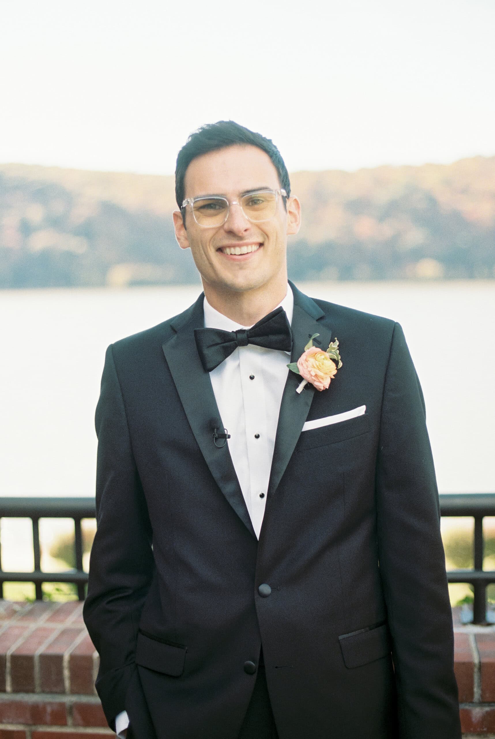 Groom Matt smiling in black tuxedo at The Grandview terrace wall with Hudson River and fall hills behind him — Poughkeepsie NY