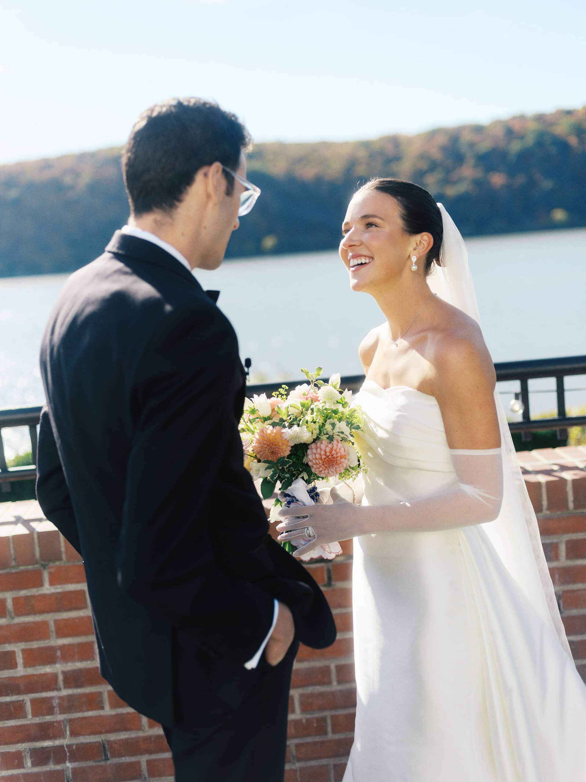 Bride Elizabeth laughing at groom Matt at The Grandview brick terrace wall with Hudson River and fall foliage — Hudson Valley wedding