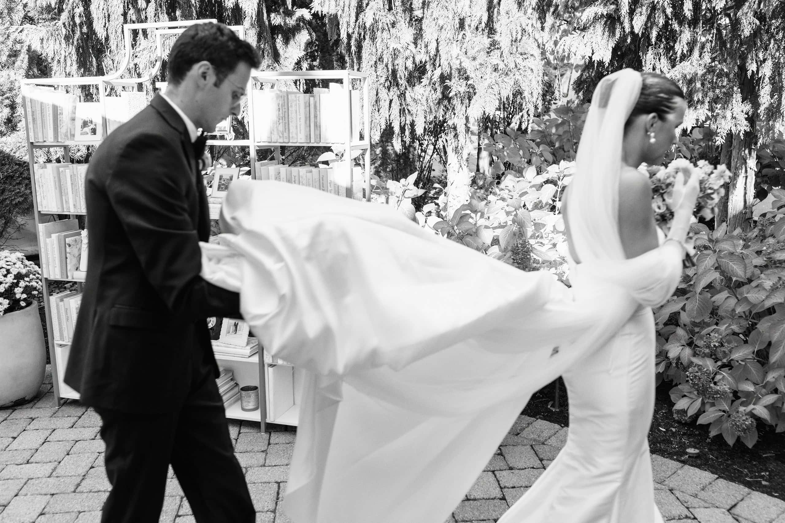 Black and white photo of Matt helping Elizabeth with her train as they walk through the garden at The Grandview — candid wedding moment