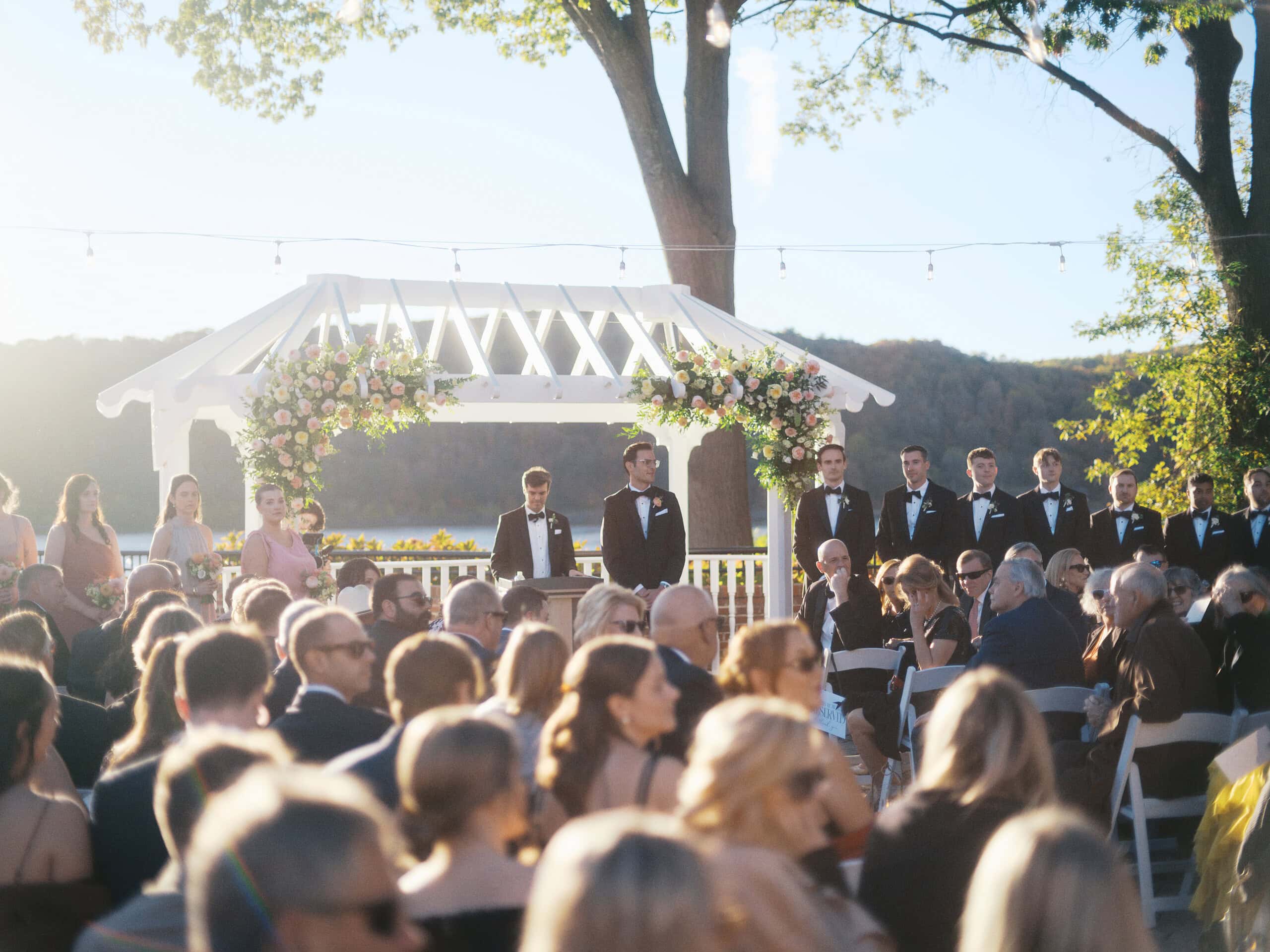 Wide view of wedding ceremony at The Grandview Poughkeepsie NY — guests seated, groom and groomsmen at floral arch with Hudson River at golden hour