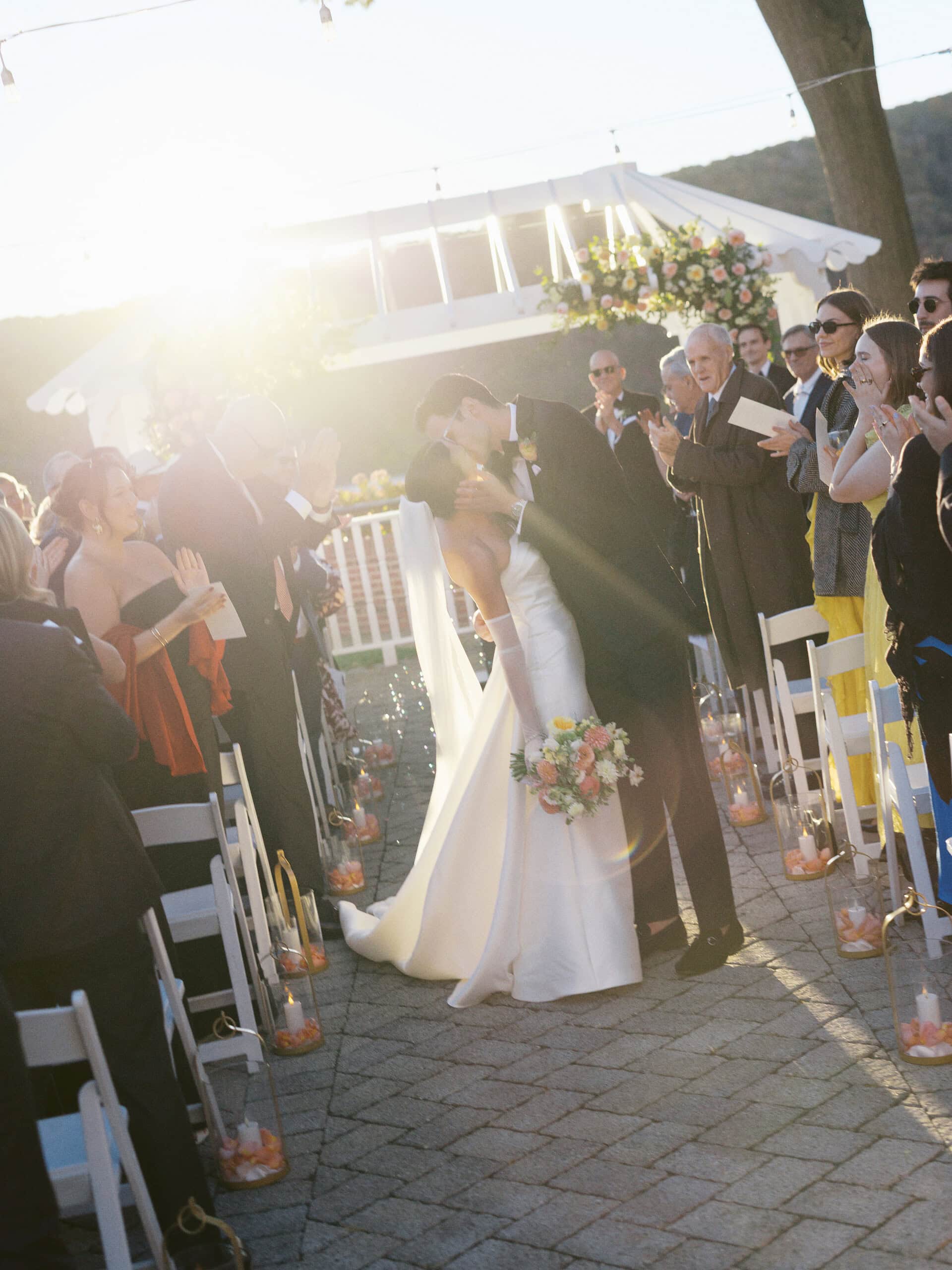 Elizabeth and Matt first kiss as newlyweds walking back down the aisle at The Grandview — backlit with golden sun flare, guests cheering
