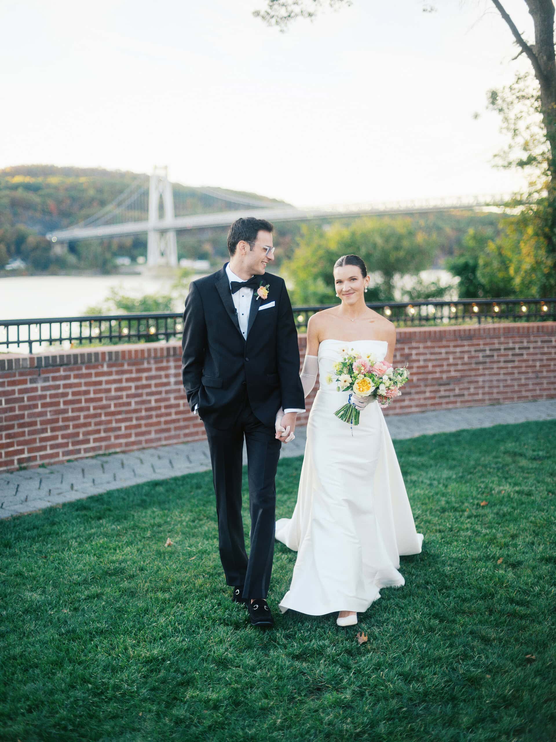 Elizabeth and Matt walking hand in hand on the lawn at The Grandview in Poughkeepsie NY with the Mid-Hudson Bridge and fall foliage behind them — Hudson Valley wedding photographer indie Bird