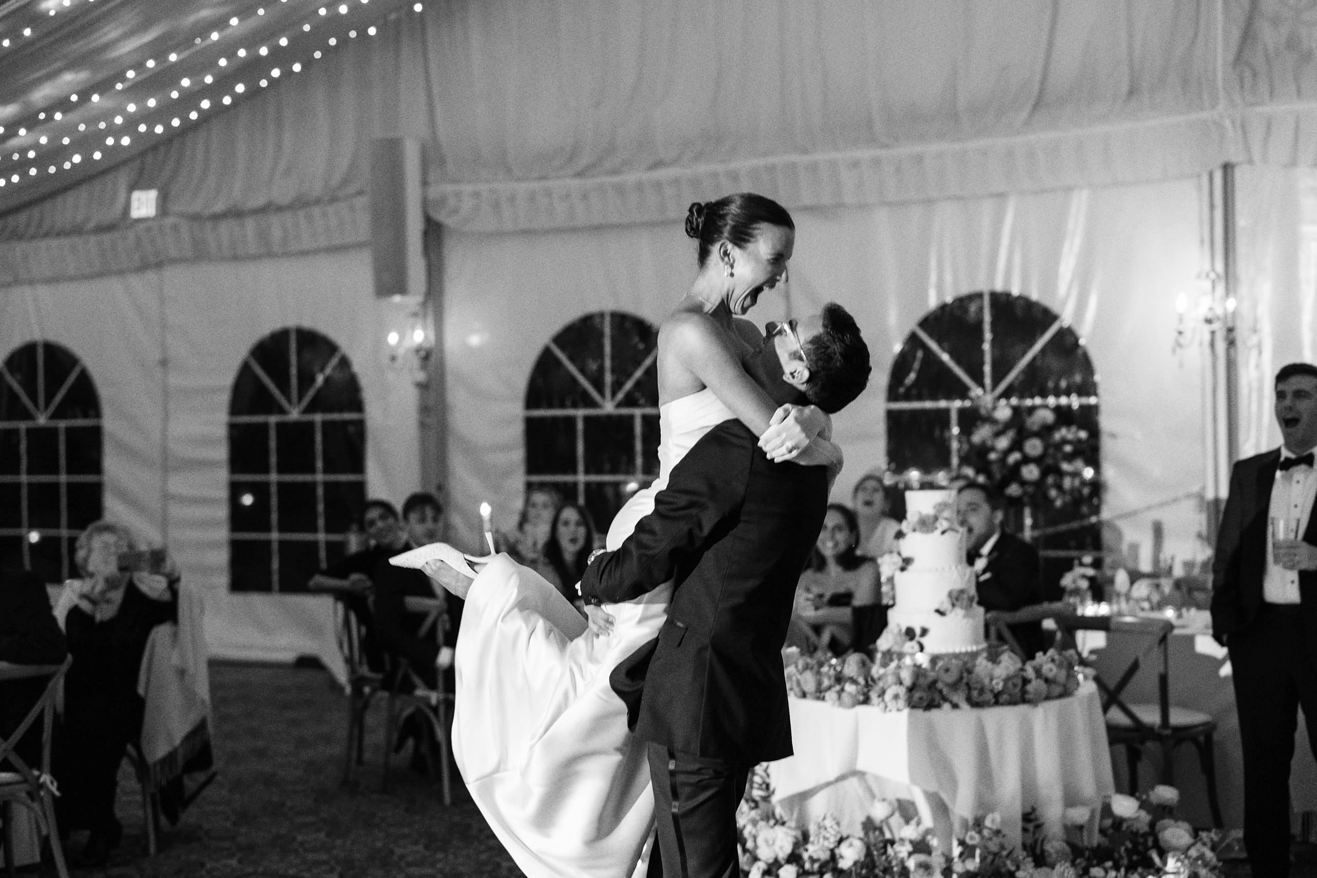 Black and white photo of Matt lifting Elizabeth during their first dance at The Grandview reception — she is laughing with joy, fairy lights overhead
